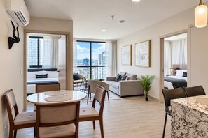 A modern hotel room with a dining area, a sofa, a bed, and a view of the cityscape. - 2BR Costa del Este Condo | City & Sea Views (Panamá)