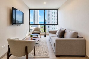 A modern living room with a sofa, armchair, and coffee table, a flat-screen TV mounted on the wall, and a large window offering a view of high-rise buildings and the ocean. - Costa del Este 2BR Apartment | Pool & City Views (Panamá)