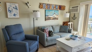 Una sala de estar con un sillón azul, un sofá gris y una mesa de centro blanca con un cuenco. Hay decoraciones en la pared, incluyendo un cuadro enmarcado titulado 'The Rocks' y un letrero que dice 'BEACH'.