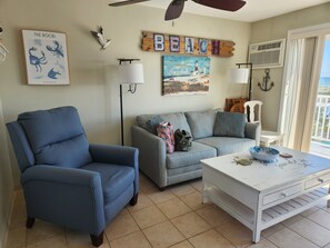 A living room with a blue armchair, grey sofa, and a white coffee table with a bowl. There are wall decorations including a framed picture titled 'The Rocks' and a 'BEACH' sign. - Oceanfront 1-bedroom condo on North Beach with a pool! (Hampton)