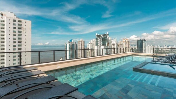 A rooftop pool with lounge chairs, a city skyline view, and high-rise buildings in the background.