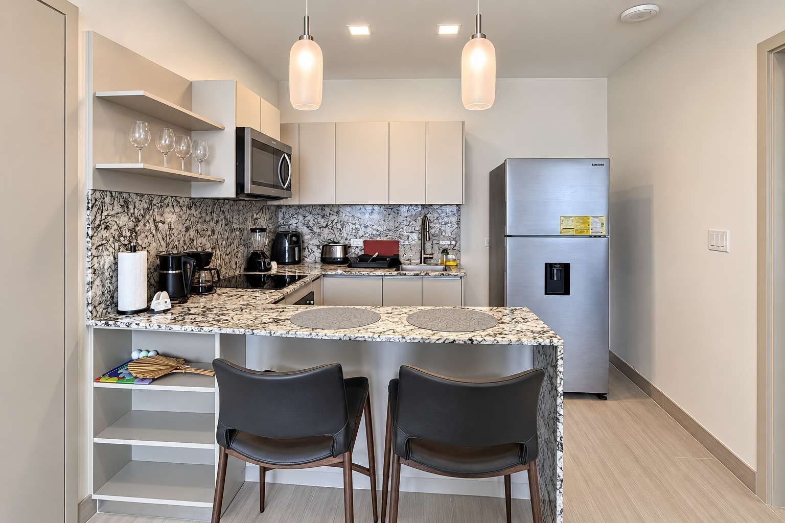 A compact kitchen with a granite countertop, stainless steel appliances, and a refrigerator. There are two bar stools and a small island with a sink.
