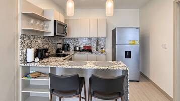 A compact kitchen with a granite countertop, stainless steel appliances, and a refrigerator. There are two bar stools and a small island with a sink.