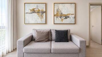 A neutral-toned sofa with two cushions, a large window with sheer curtains, and two abstract paintings on the wall.