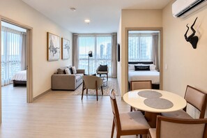 A modern hotel room with a bed, a sofa, a dining table with chairs, and a city view. - 2BR Costa del Este Condo | Pool, Gym & Sauna (Panamá)