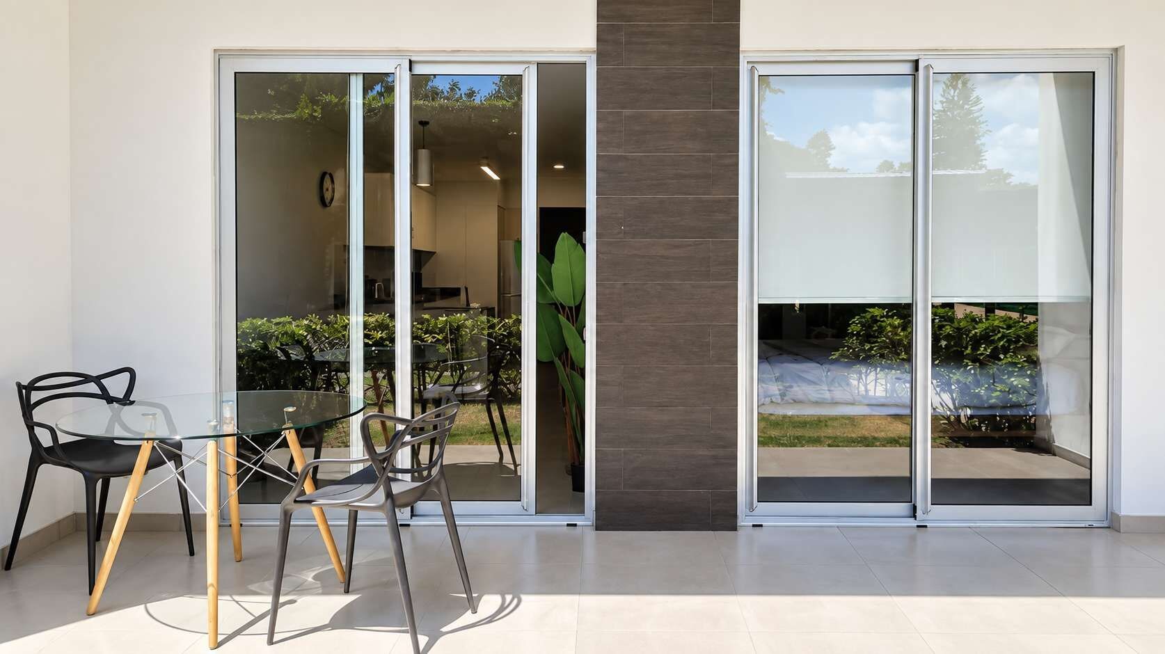 Un espace de restauration extérieur moderne, doté d’une table et de chaises en verre, d’un pilier en bois et offrant une vue sur un jardin grâce à des portes-fenêtres coulissantes.