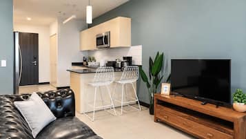 Una sala de estar moderna con un sofá de cuero, un mueble bajo para televisor de madera y una zona de cocina con microondas y taburetes de bar.