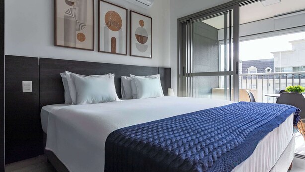 Een moderne hotelkamer met een groot bed, een donker hoofdeinde en uitzicht op de stad door grote ramen.