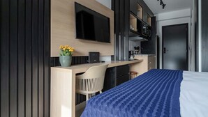 Een moderne hotelkamer met een bed, een bureau met stoel, een flatscreen televisie, een kleine potplant en een kitchenette.