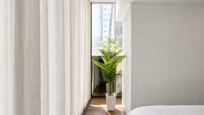 Una camera d'hotel moderna con un letto grande, tende bianche, una pianta in vaso e vista su un edificio.