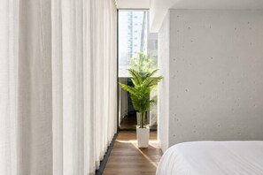 Ein modernes Hotelzimmer mit einem großen Bett, weißen Vorhängen, einer Topfpflanze und Blick auf ein Gebäude durch das Fenster.