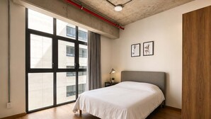A bedroom with a large window, a bed, a nightstand, and two framed pictures on the wall.