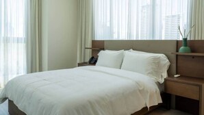 Ein Hotelzimmer mit einem Bett, einem Nachttisch, einem Telefon und Blick auf Gebäude durch transparente Vorhänge.