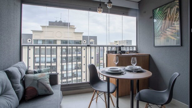 A modern living room with a balcony, a dining area, and a view of buildings. - Furnished Studio in São Paulo with City Views (São Paulo)