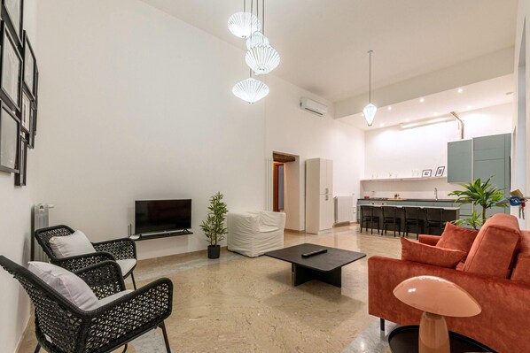 Uma sala de estar moderna com televisão de tela plana, cadeiras de vime preto, sofá vermelho, mesa de centro e área de cozinha com banquetas.