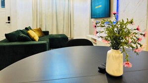Ein modernes Wohnzimmer mit einer Couch, einem runden Tisch, einer Vase mit Blumen, einem an der Wand befestigten Flachbildfernseher und einem Fenster mit Vorhängen.