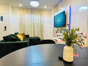 Ein modernes Wohnzimmer mit einer Couch, einem runden Tisch, einer Vase mit Blumen, einem an der Wand befestigten Flachbildfernseher und einem Fenster mit Vorhängen.