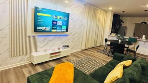 Ein modernes Wohnzimmer mit Flachbildfernseher, einem grünen Sofa, einem gemusterten Teppich und einer Kristallleuchter.