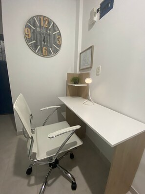 Un espacio de oficina moderno con un escritorio blanco, una silla de oficina blanca, un reloj de pared, un estante pequeño con una planta y un cuadro enmarcado.