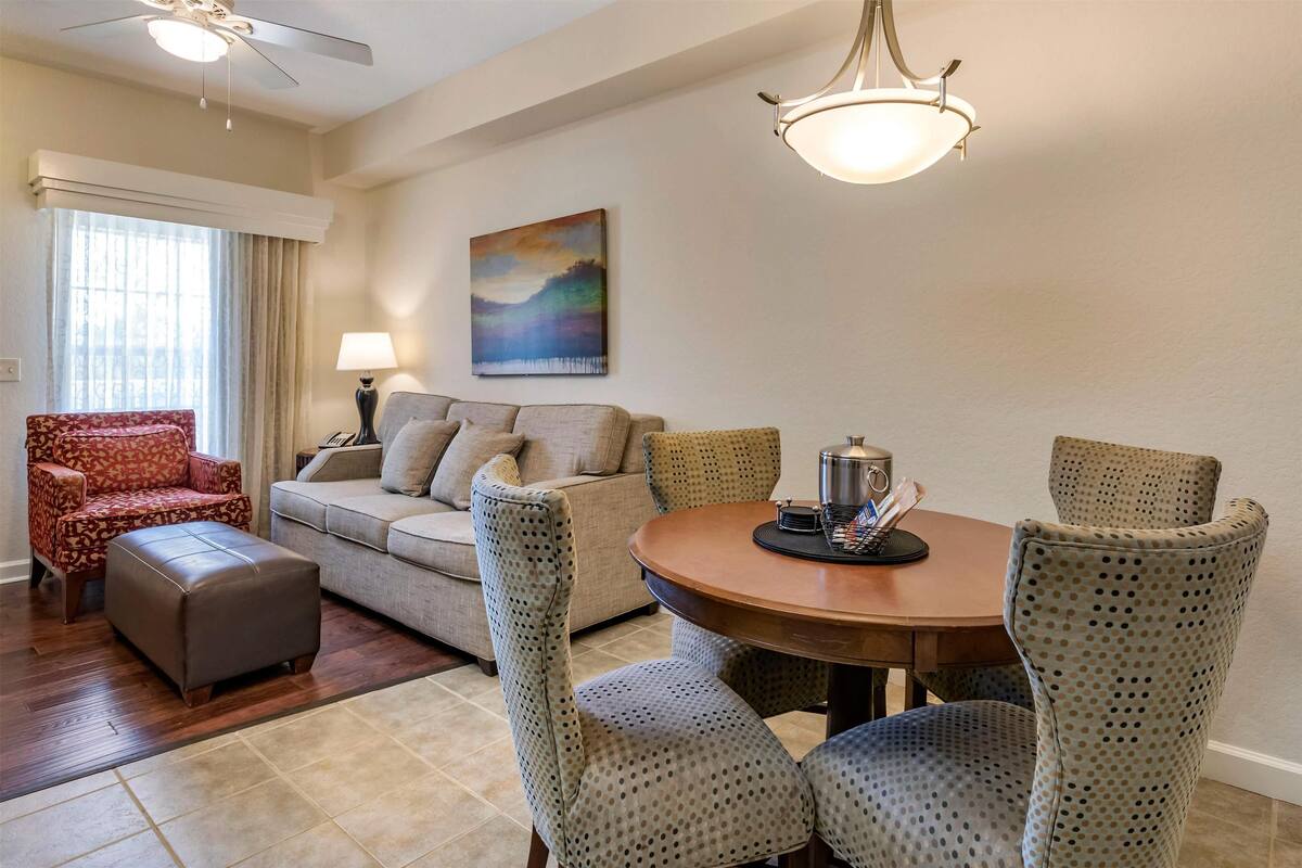 Un salon comprenant un canapé, un pouf et des fauteuils à motifs disposés autour d’une table ronde, sur laquelle se trouve une lampe, et un tableau accroché au mur.