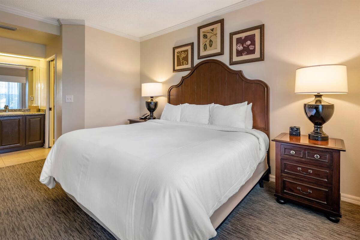 Une chambre d’hôtel avec un grand lit, une table de chevet avec une lampe, une commode et des œuvres d’art encadrées accrochées au mur.