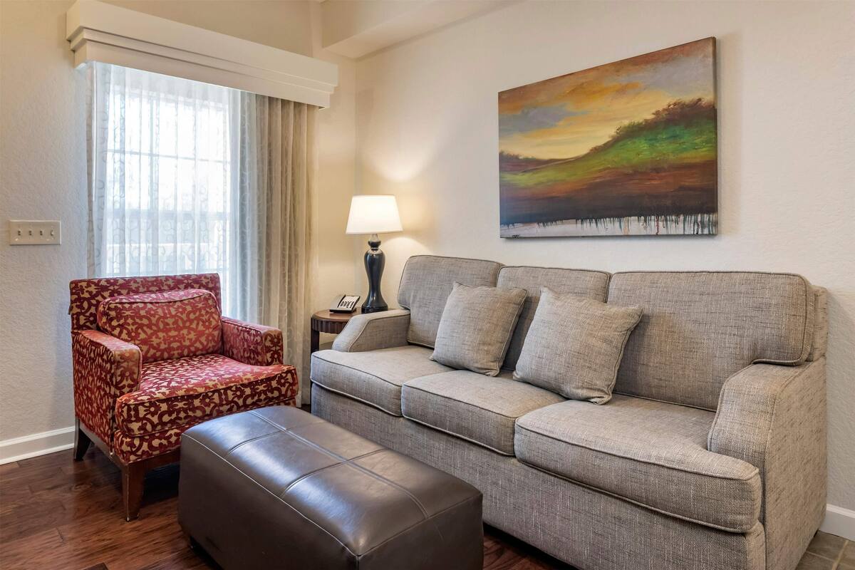 Un salon avec un fauteuil à motifs, un canapé gris, un pouf marron, une lampe et un tableau accroché au mur.