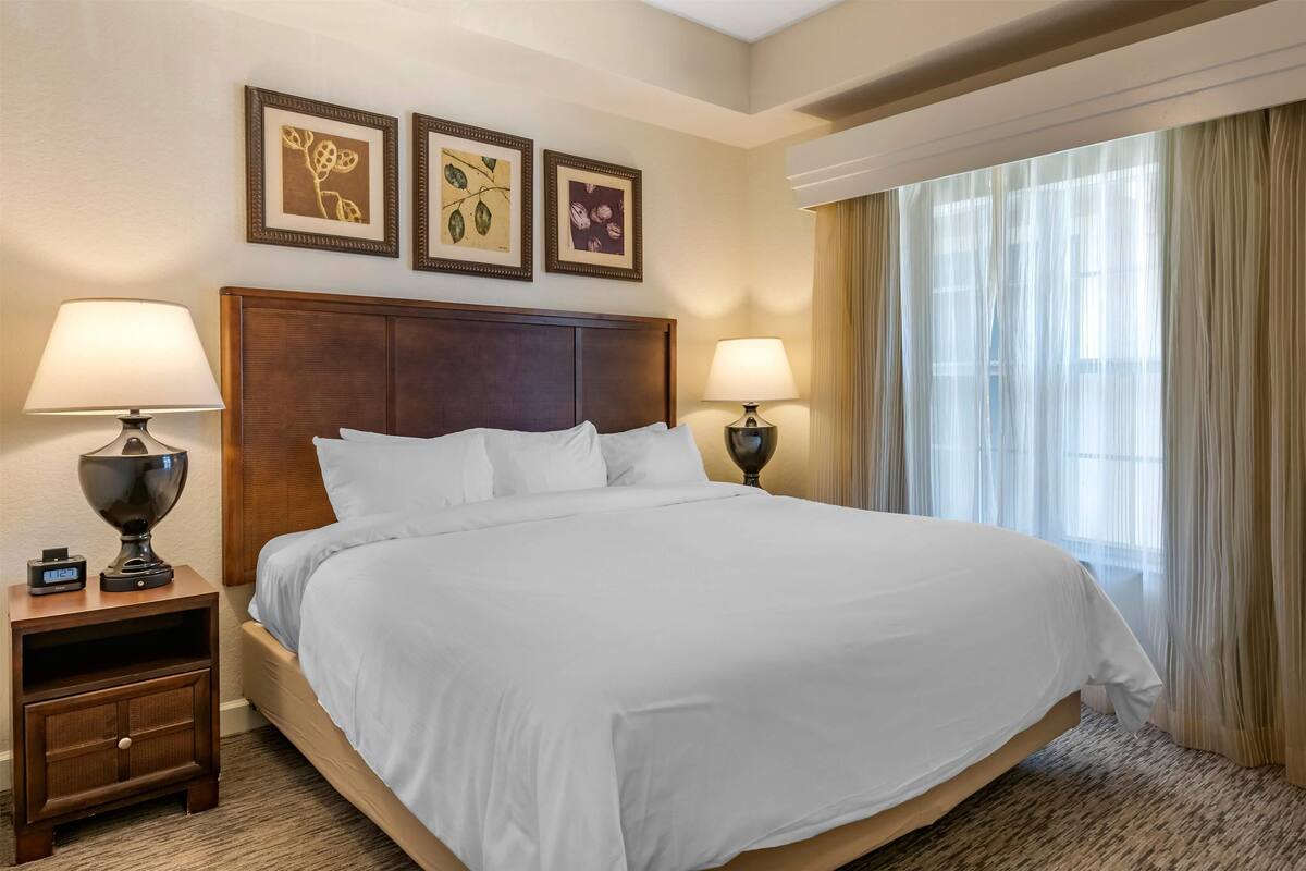 Une chambre d’hôtel avec un grand lit, deux tables de chevet avec des lampes, une horloge et trois tableaux encadrés accrochés au mur.