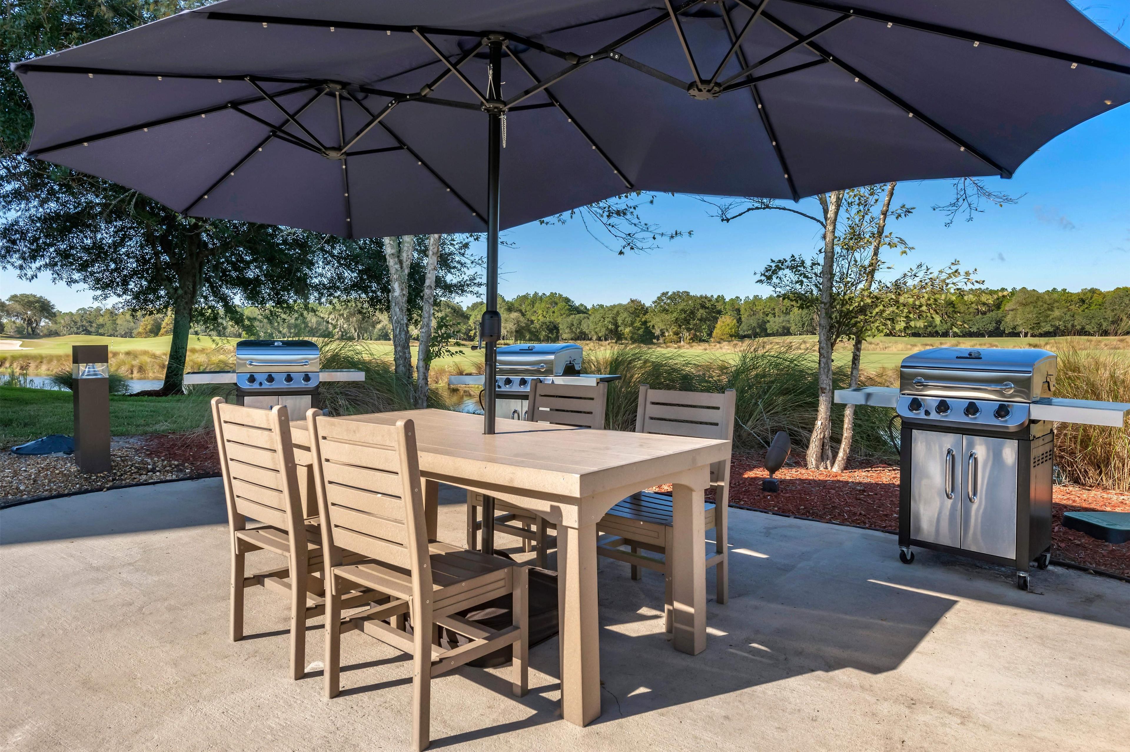 An outdoor patio with a table and chairs, a large umbrella, and two barbecue grills.