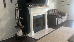 Un salon avec une cheminée, un canapé, un téléviseur fixé au-dessus de la cheminée et une étagère décorative agrémentée de plantes.