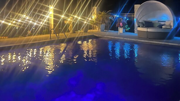 Une scène nocturne avec une piscine reflétant les lumières, un espace de repos abrité et une structure illuminée par des faisceaux de lumière.