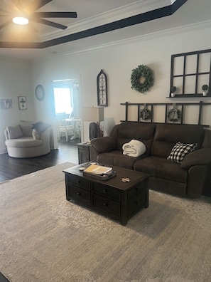 A living room with a dark brown sofa, a coffee table, a white armchair, and a dining area in the background. - By The Pool (Winchester)