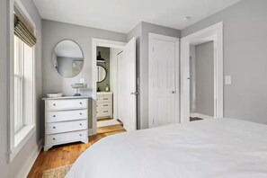 A bedroom with a large bed, a dresser, a mirror, and a door leading to a bathroom. - Charming Home with Hot tub & walk to town! (Three Oaks)