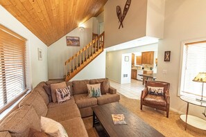 A living room with a sectional sofa, a coffee table, a brown leather armchair, a wooden staircase, and a kitchen in the background. - Located near SHARC, walking trails, & eateries, HT (Sunriver)