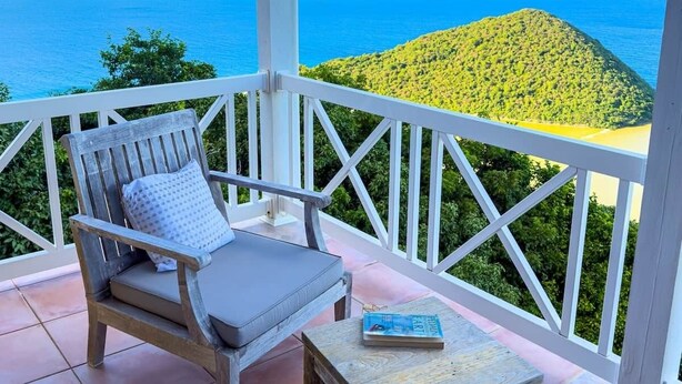 A balcony with a chair and a small table, overlooking a scenic view of the sea and a green hill.