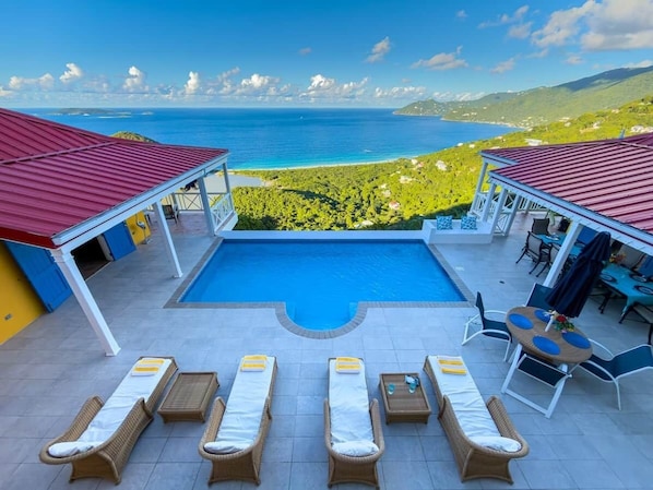 A pool area with lounge chairs, a dining table, and a view of the ocean and hills.