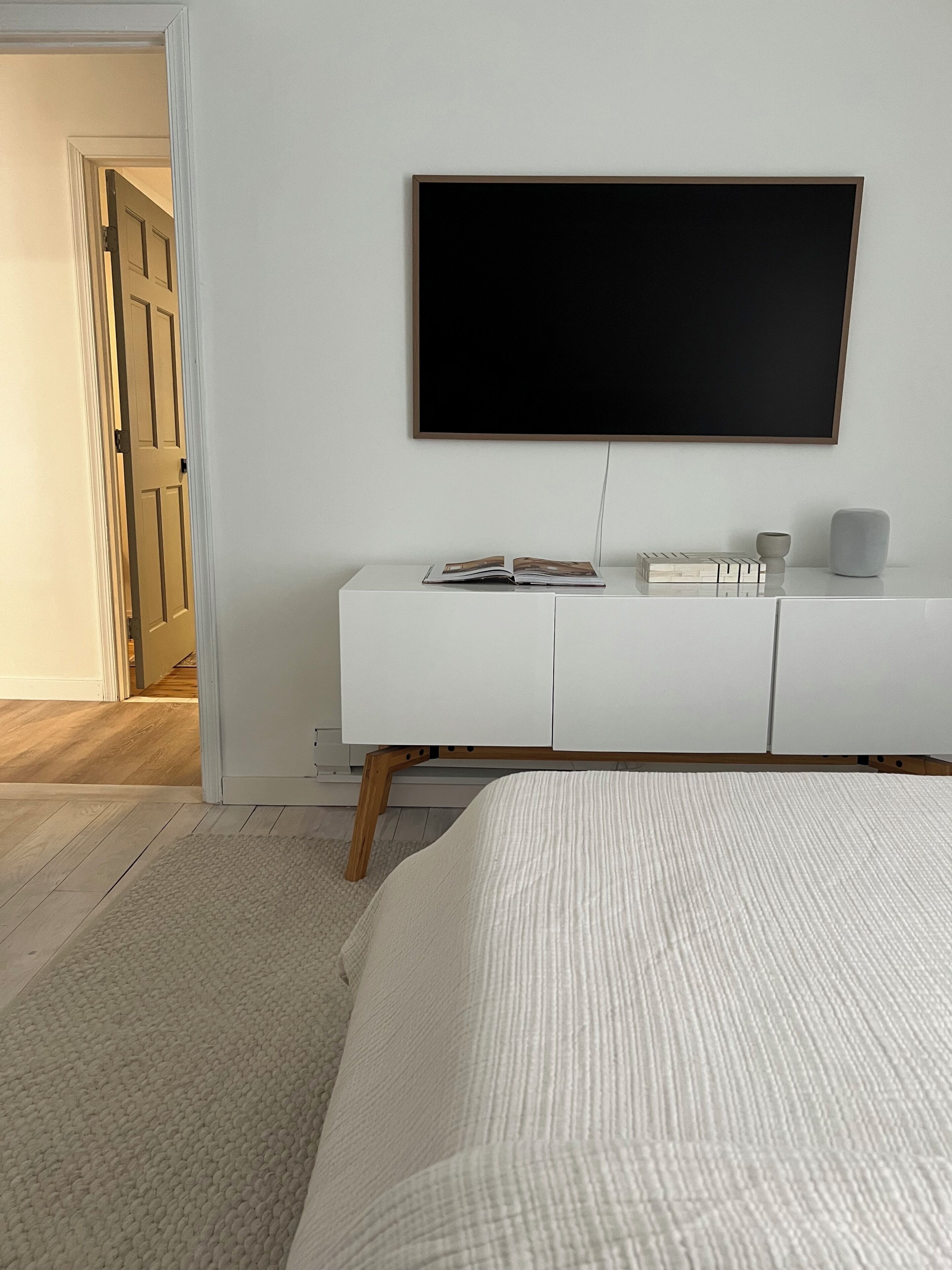 A bedroom with a bed, a white dresser, a flat-screen TV, and a door leading to another room.