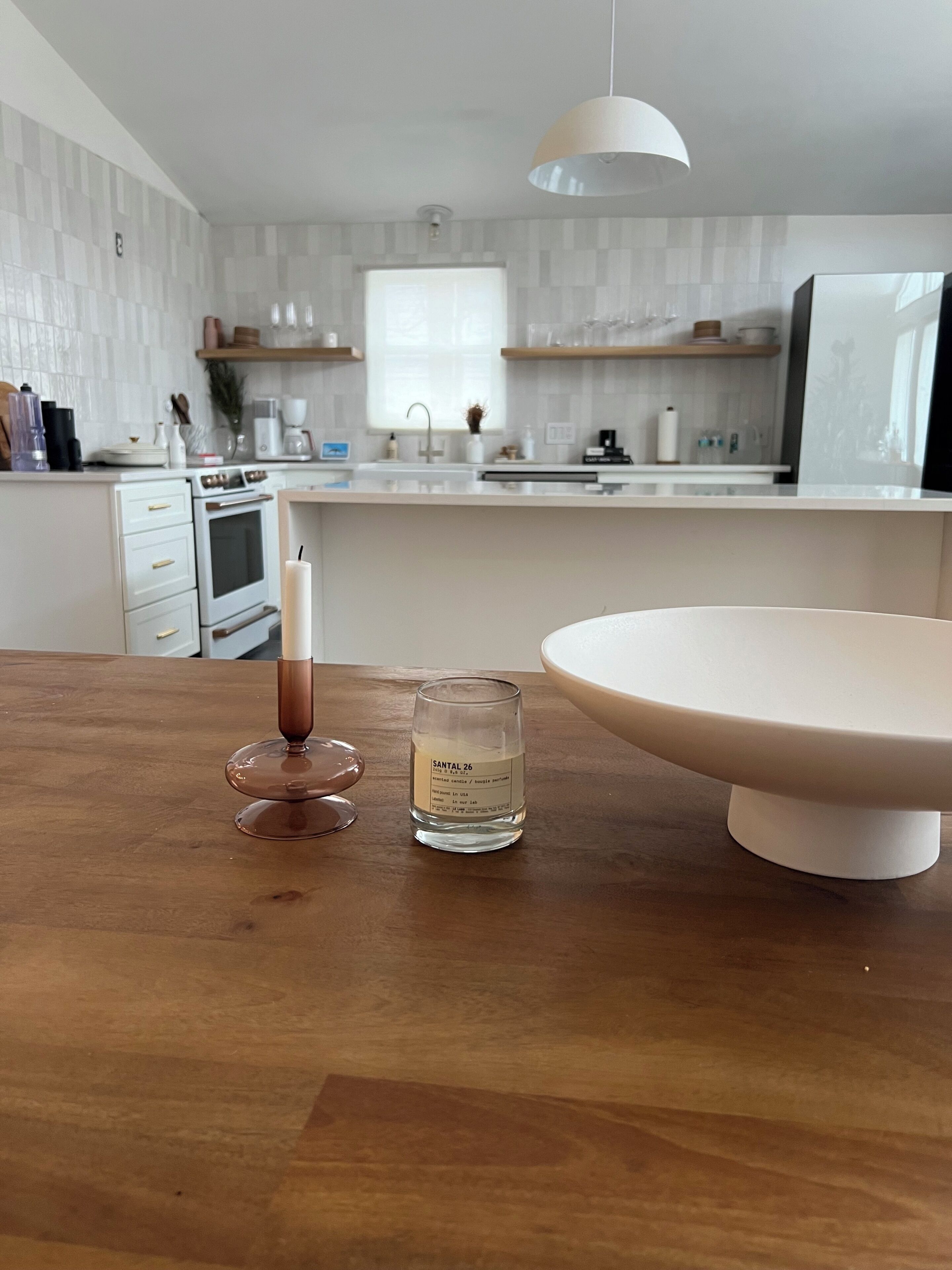 A kitchen with white cabinets, a white countertop, a hanging light, and a wooden table with a candle and a glass of water.