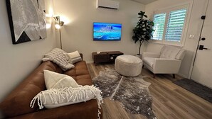 A modern living room with a brown leather sofa, a white armchair, a round ottoman, a large painting, and a potted plant. - Cozy 1-bedroom condo in enjoyable Chico with AC (Chico)