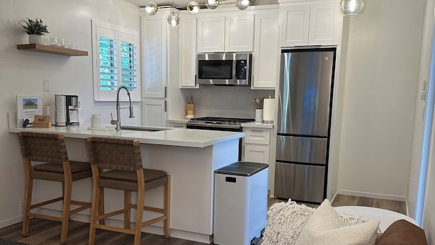 A modern kitchen with white cabinets, a stainless steel refrigerator, and a built-in microwave. There is a small island with a sink and two bar stools. - Cozy 1-bedroom condo in enjoyable Chico with AC (Chico)