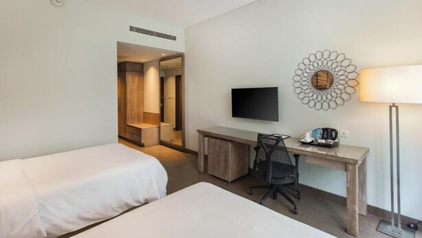 A hotel room with two beds, a desk with a chair, a TV, a lamp, and a decorative wall piece. - PALMERAS DEL CARIBE - STUDIOS and SUITES (Punta Cana)