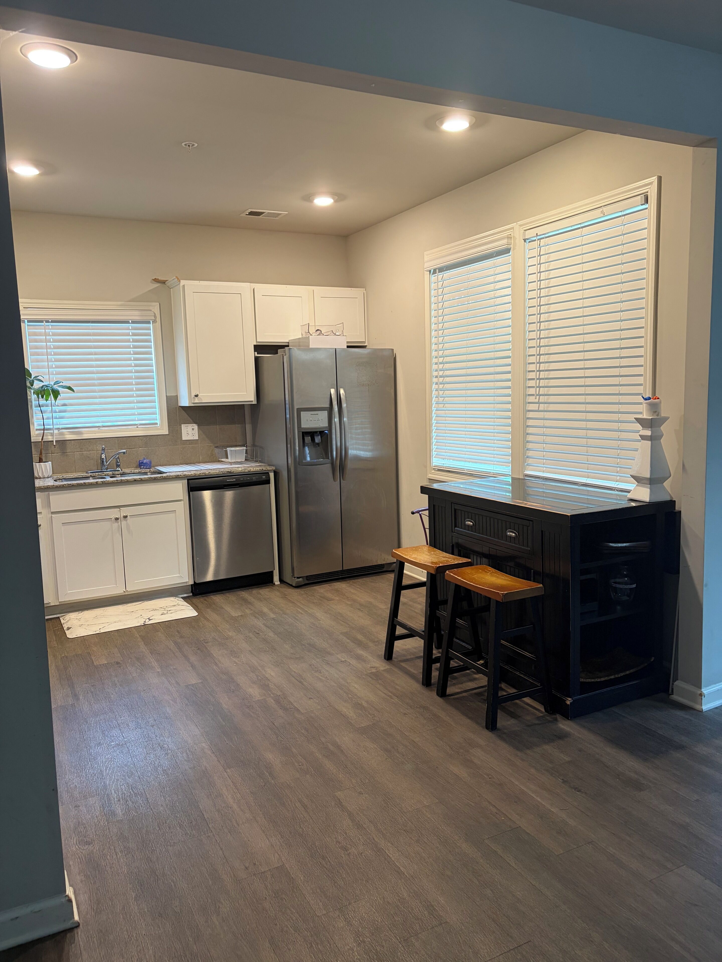 A kitchen with a stainless steel refrigerator, white cabinets, a wooden bar stool, and a window with blinds.