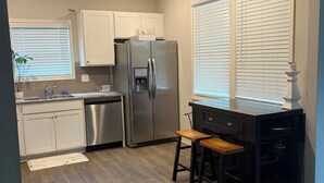Una cocina con refrigerador de acero inoxidable, armarios blancos, un taburete de bar de madera y una ventana con persianas.