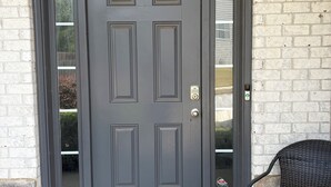 Puerta principal de color gris oscuro con un panel de vidrio, felpudo con huellas de patas y una silla de mimbre en el exterior.