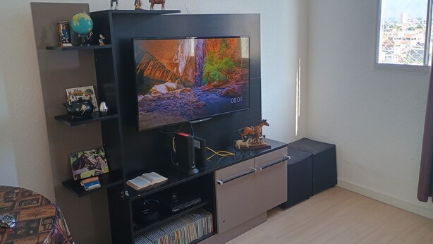 A living room with a flat-screen TV, shelves with decorative items, a coffee table with a patterned table runner, and a window with a city view. - O melhor de Madureira  (Rio de Janeiro)