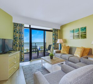 A modern living room with a sofa, coffee table, TV, and a balcony with a view. - 3BR Oceanfront Escape w/ Panoramic Views! (Myrtle Beach)