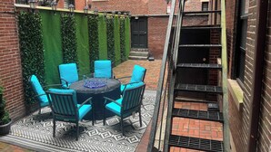 Un cortile con un tavolo rotondo e sedie, un edificio in mattoni, un muro verde con piante rampicanti e una scala in metallo.