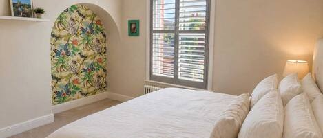 Un dormitorio con cama, ventana con persianas, radiador y papel tapiz con diseño floral.
