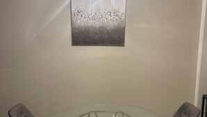 Un comedor con una mesa de vidrio, dos sillas tapizadas, un alfombra de estampado geométrico y un cuadro abstracto colgado en la pared.