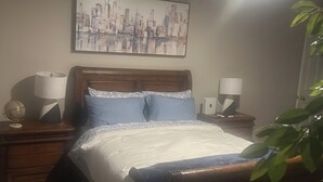 Una cama bien hecha con cabecera, dos mesitas de noche con lámparas, un cuadro de un paisaje urbano en la pared y una planta en maceta.