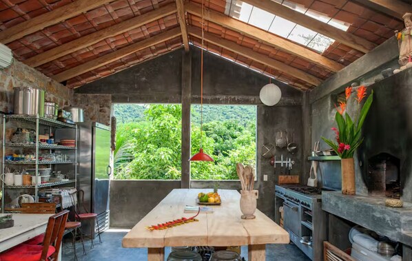 Una cocina rústica con una mesa de madera, estantes con utensilios de cocina, una estufa y vistas a vegetación exuberante.